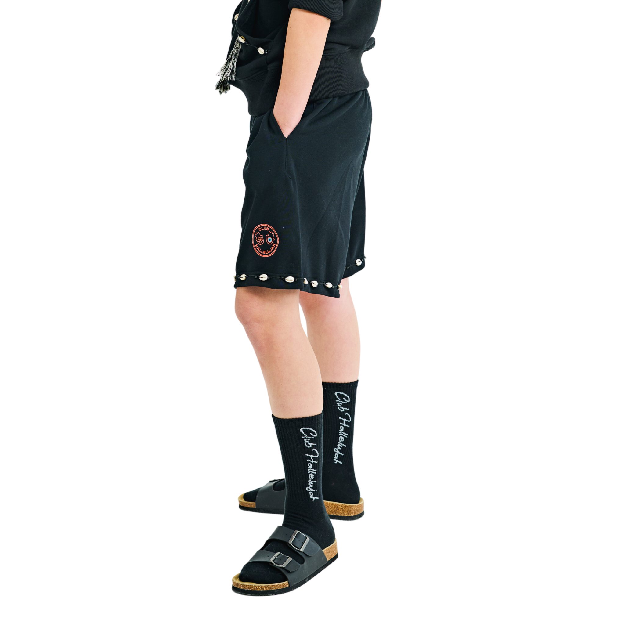 Side view of female model in unisex black shorts, braided drawstring, cowrie shell hem, 100% cotton, embroidered details.