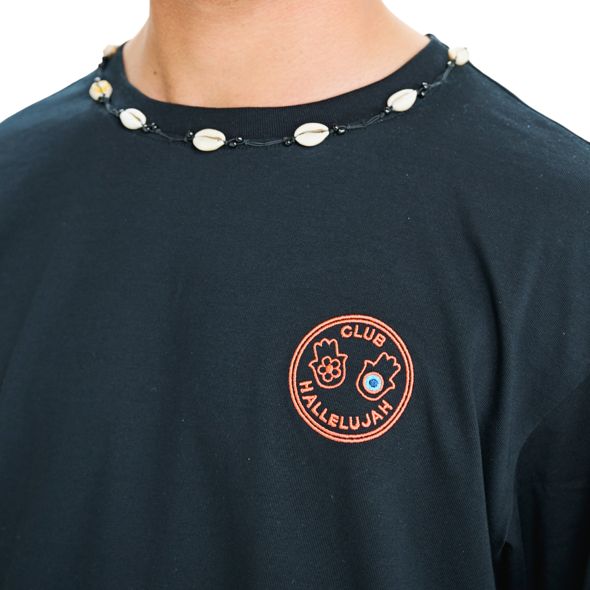 Close-up of black oversized T-shirt showing hand-embroidered 'Club Hallelujah' front logo patch and cowrie shell neckline