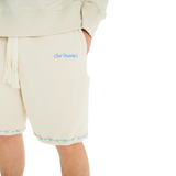 Close-up of unisex stone shorts, Club Hallelujah embroidered logo, handmade braided drawstring, cowrie shell trim with blue thread detailing at hem